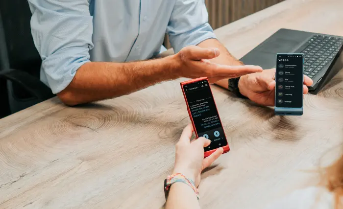 Two people sit at a wooden table, each holding a translator with apps open on the screens. One person gestures toward the other, and a keyboard is visible in the background.